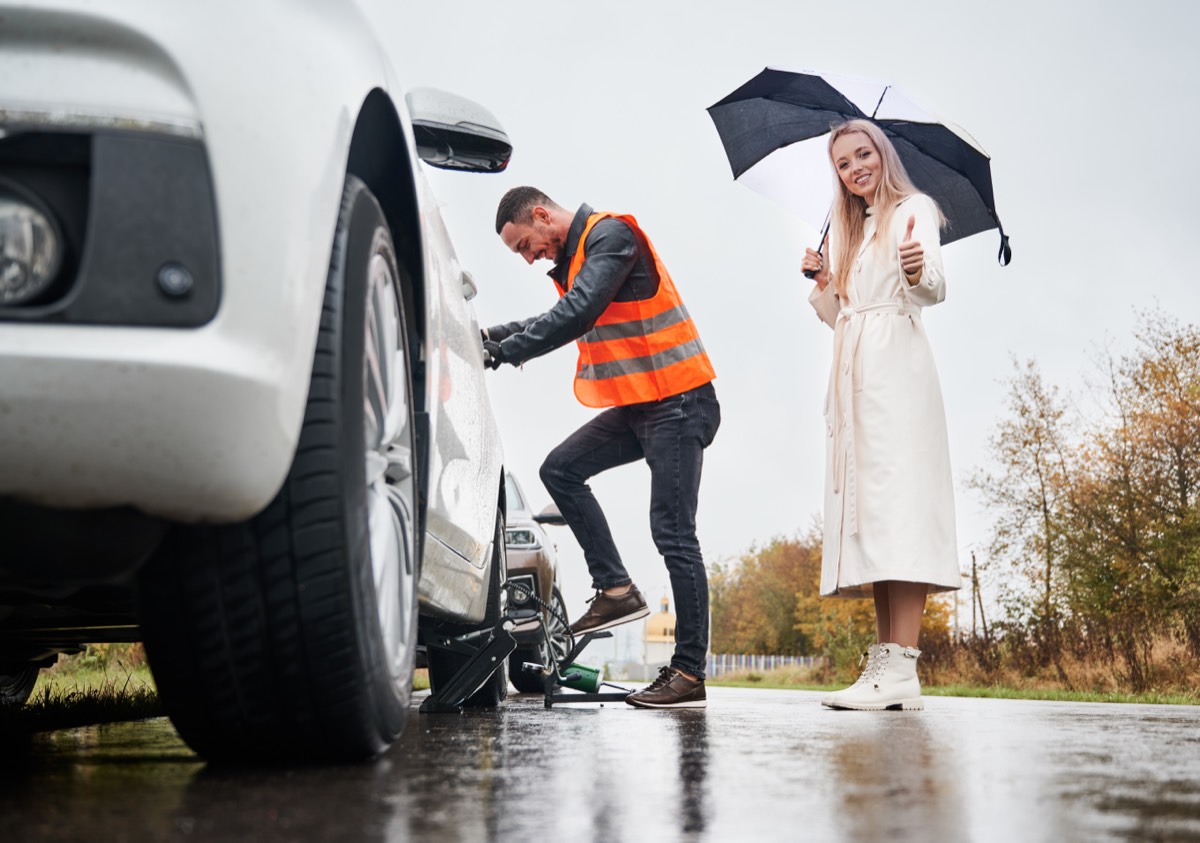 Pannenhilfe im Regen – Mechaniker in Warnweste wechselt Reifen, Autofahrerin mit Schirm steht daneben