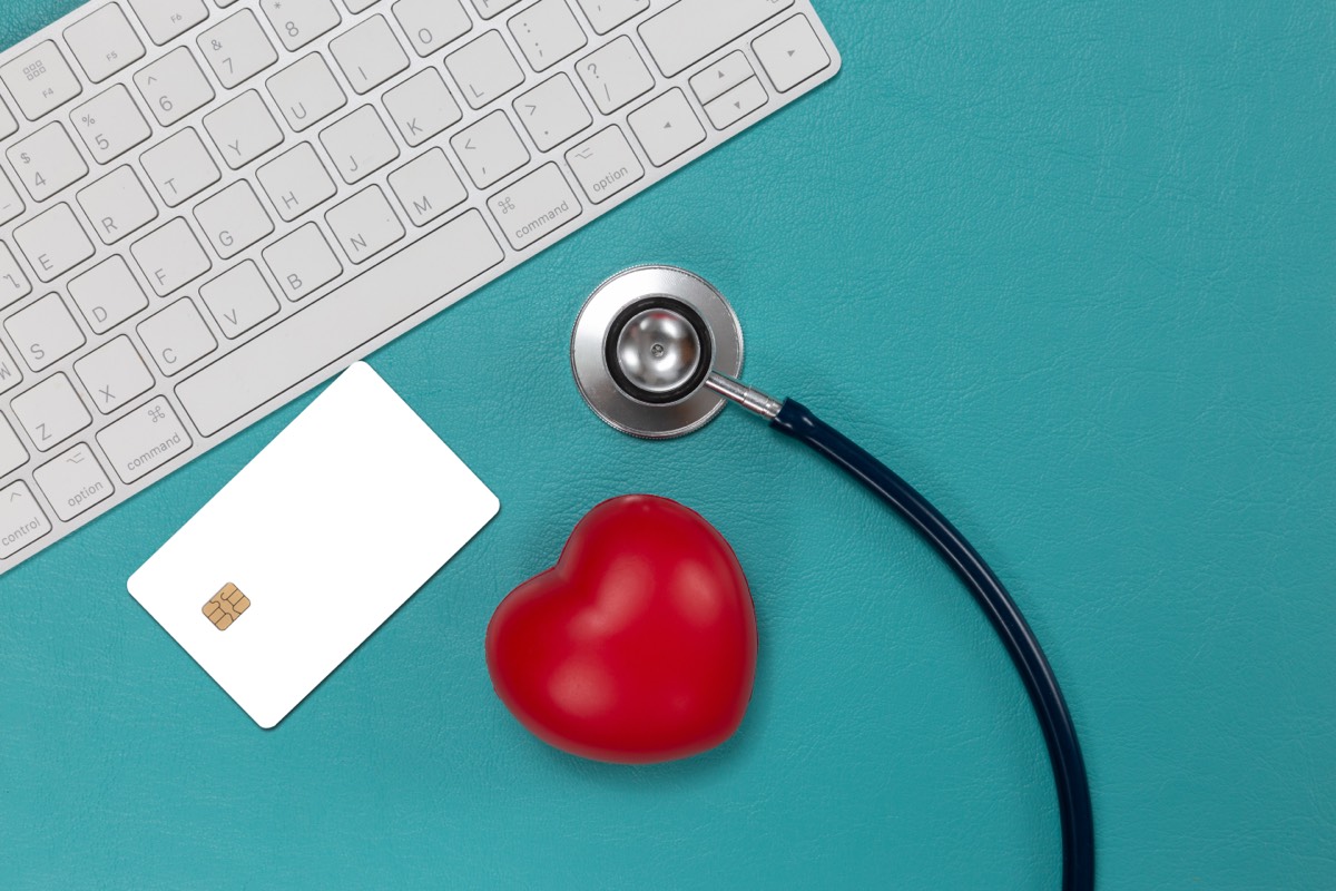 Draufsicht auf eine weiße Chipkarte (Krankenversicherungskarte), ein Stethoskop und ein rotes Herz auf türkisfarbenem Untergrund neben einer weißen Tastatur