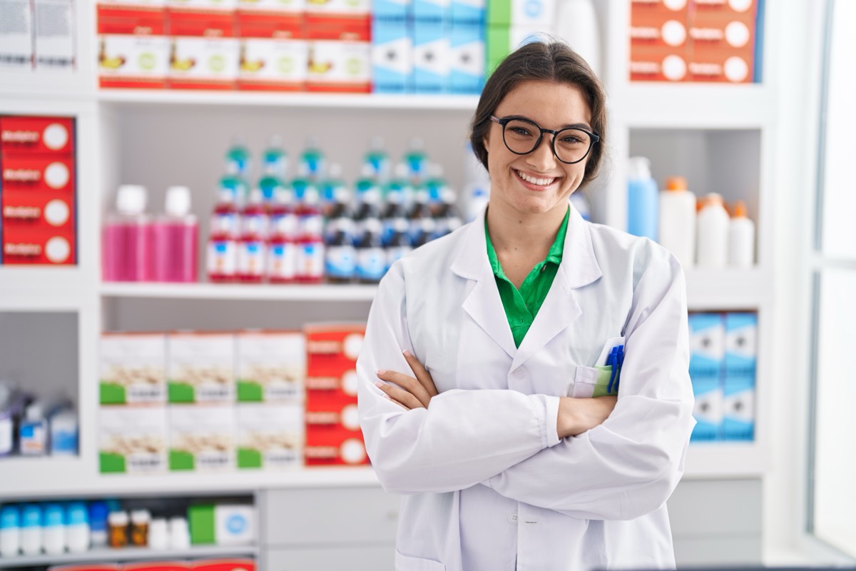 Lächelnde Apothekerin mit Brille und weißem Kittel steht mit verschränkten Armen vor gut sortierten Regalen einer Farmacia