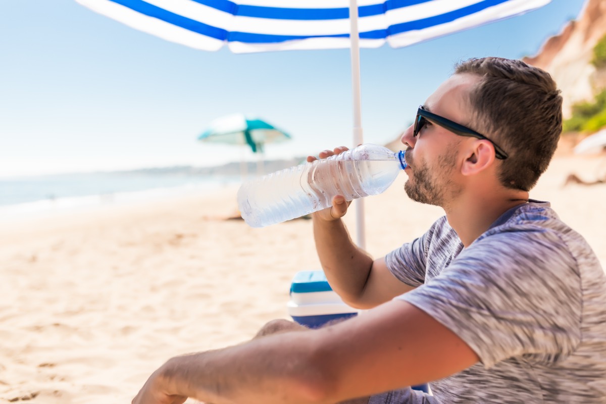 Mann am Strand trinkt Wasser unter Sonnenschirm – Dehydrierung durch Hitze auf Lanzarote