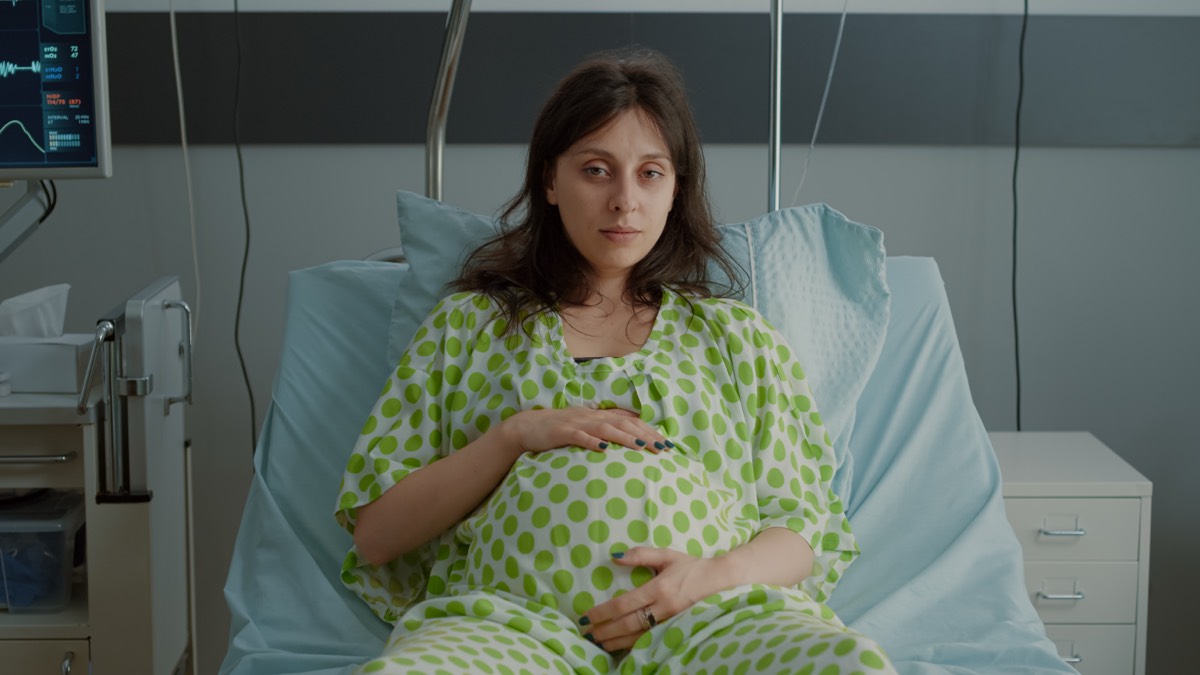 Schwangere Frau mit rundem Bauch liegt in einem Krankenhausbett und hält beide Hände auf den Bauch – medizinische Geräte und Monitore im Hintergrund, Vorbereitung auf die Entbindung