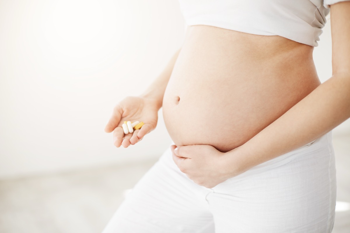 Nahaufnahme einer schwangeren Frau, die lächelnd ihren runden Bauch hält und in der anderen Hand eine Handvoll Vitamintabletten und Kapseln – Schwangerschaftsvitamine und Folsäure