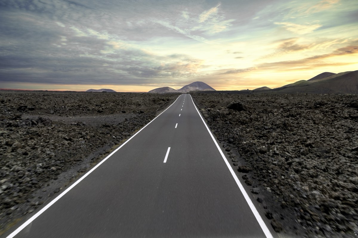 Gerade Asphaltstraße durch die schwarze Lavawüste des Timanfaya-Nationalparks auf Lanzarote – Vulkankegel am Horizont, dramatischer Abendhimmel mit goldenen Wolken – typische Straße die man mit dem Mietwagen auf Lanzarote befährt